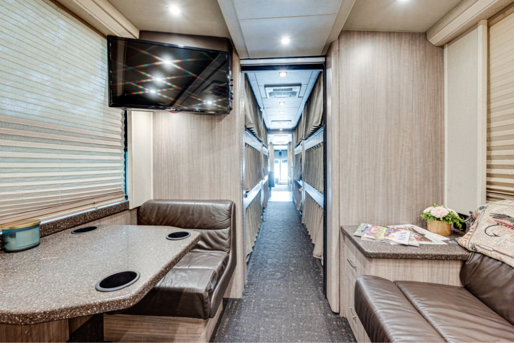 View of the dining area with booth seating and a mounted flat-screen TV inside a Prevost H345 bus, offering a comfortable space for meals or meetings during travel.