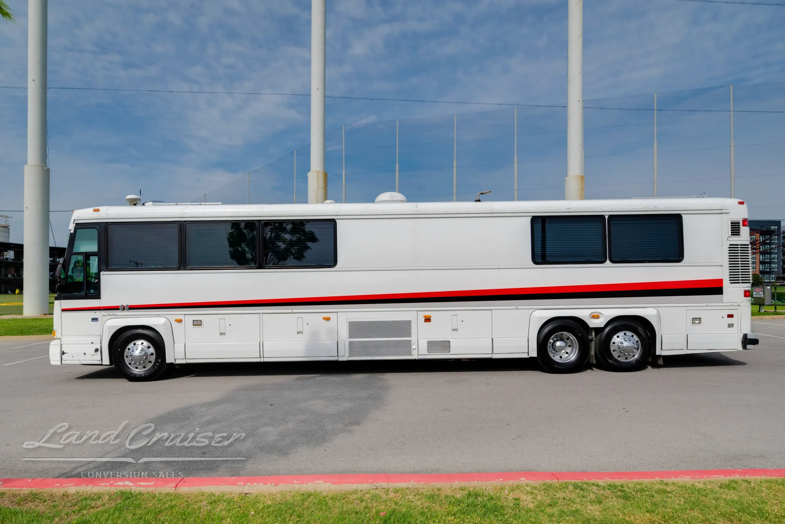 Side profile of MCI bus conversion featuring white exterior, red accent stripe, and triple axle configuration