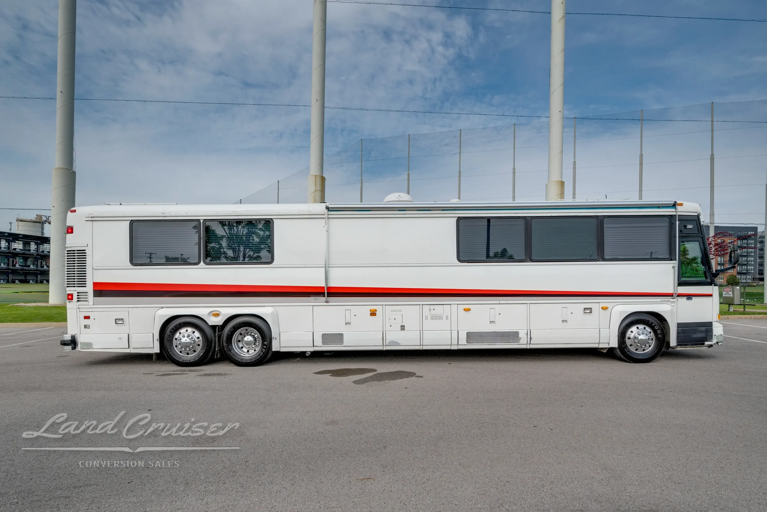 Full driver side view of 1998 MCI executive bus with red stripe, slide-out compartments, and triple axle