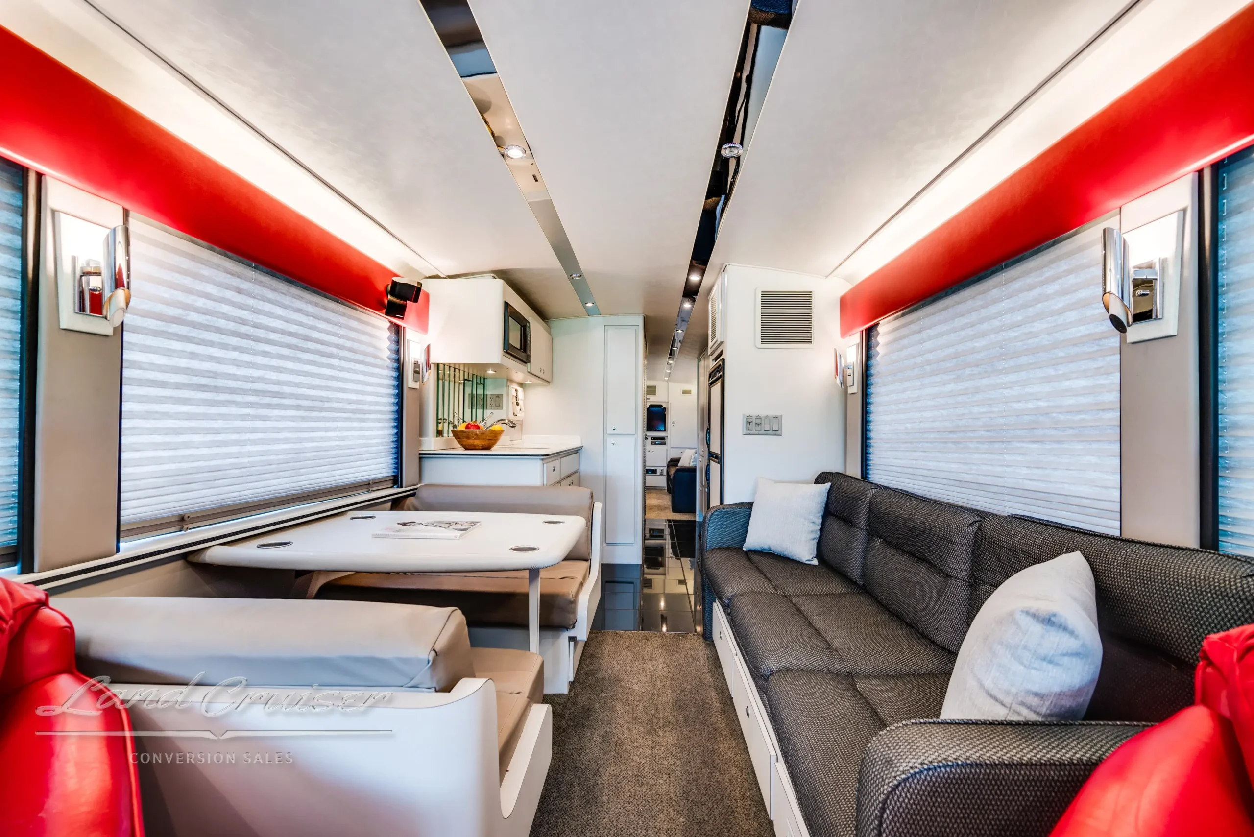Living area of MCI Executive Coach with beige booth dinette, long sleeper sofa, and red chairs