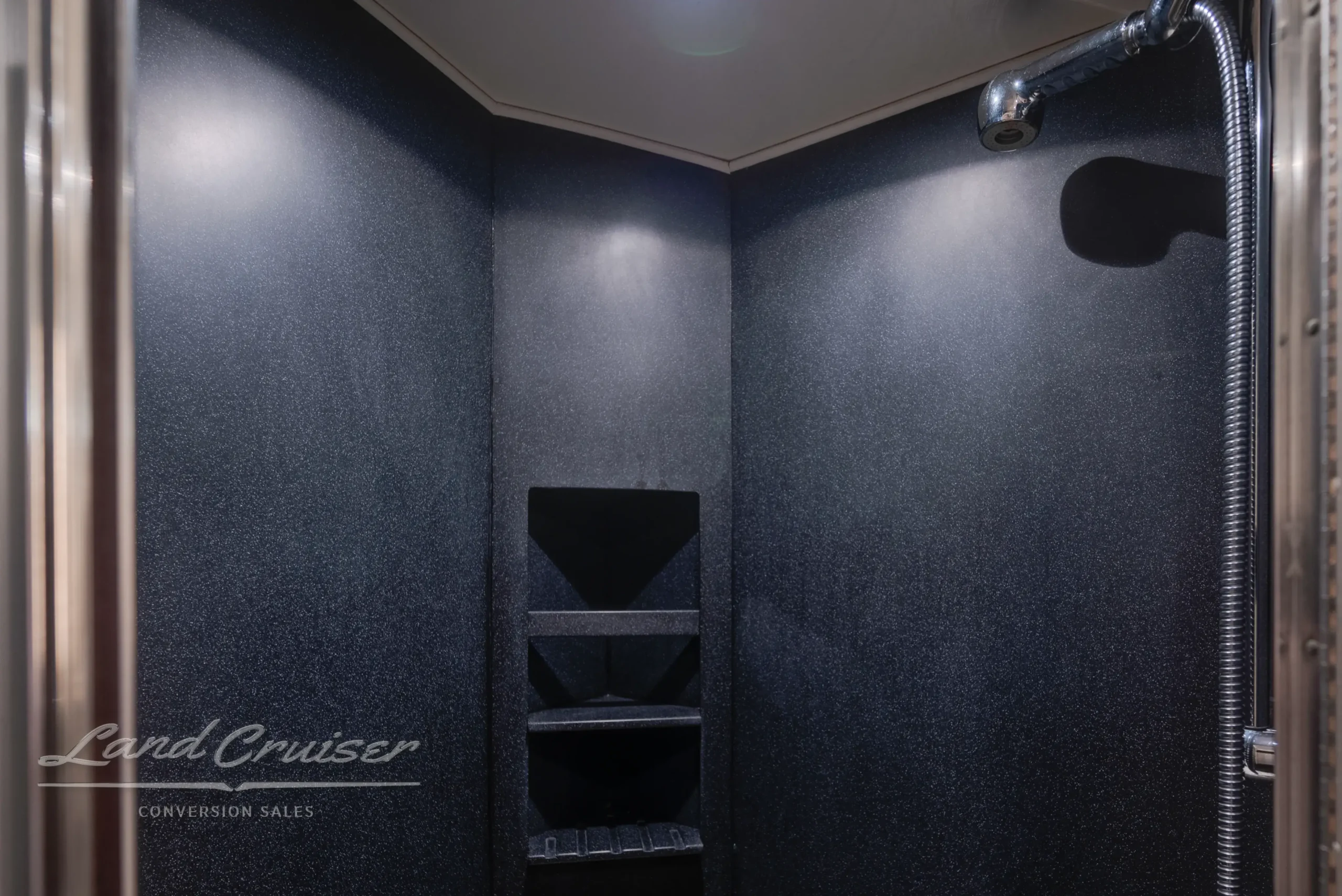 Walk-in shower inside MCI Executive Coach featuring dark tile walls, built-in shelving, and overhead lighting
